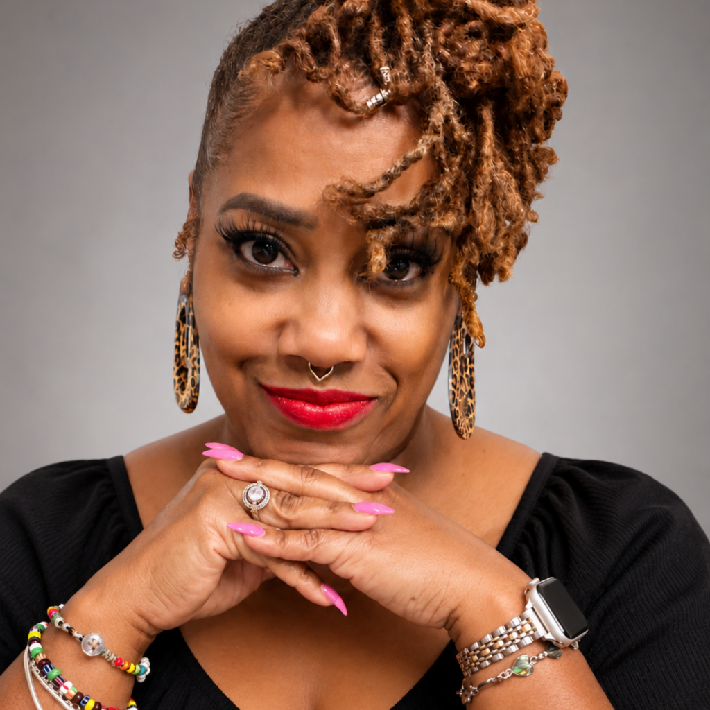 A woman with styled locs, hoop earrings, and bright pink nails smiles gently, resting her chin on her folded hands. She wears multiple bracelets, a watch, and a nose ring, against a neutral gray background.
