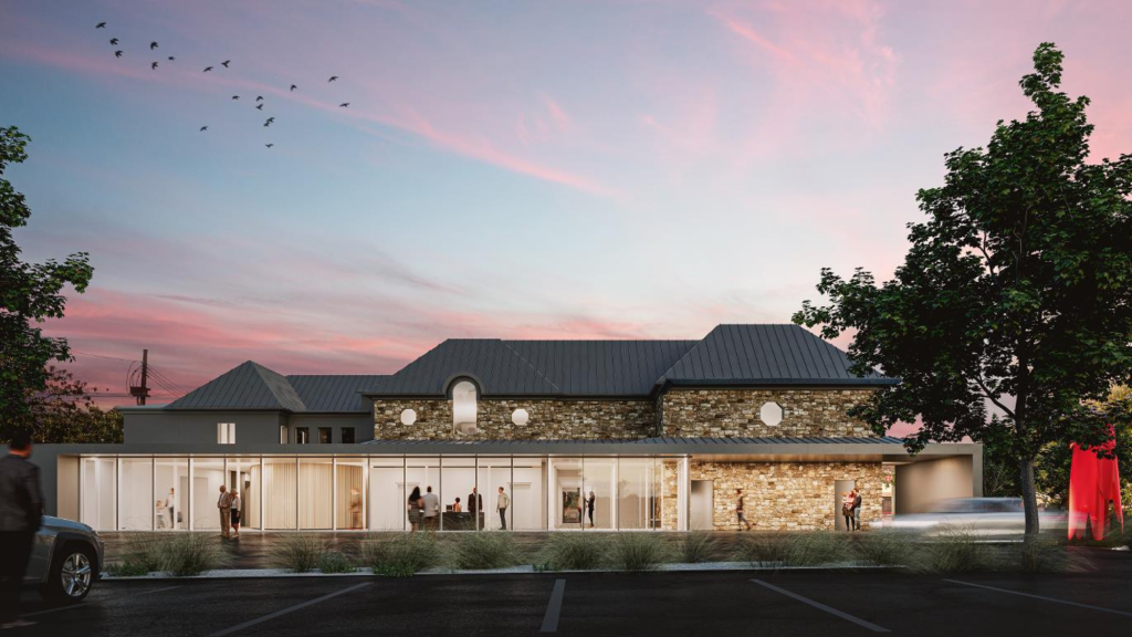 A modern building with a stone facade and large glass windows is illuminated at dusk. People are visible inside and outside. Trees and parked cars line the foreground, and birds are flying in the colorful evening sky.