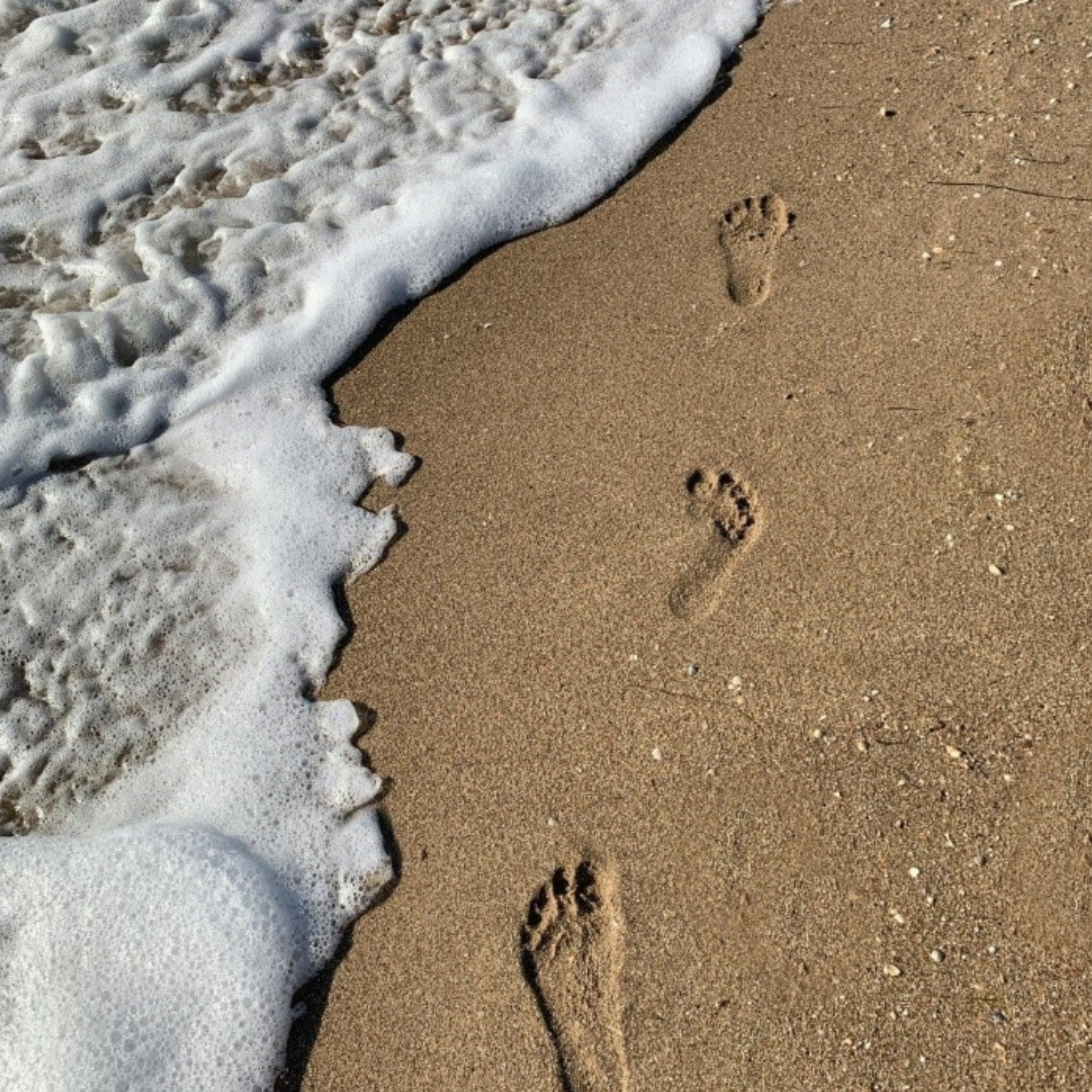 Three footprints in wet sand are about to be washed away by an incoming wave, with foam reaching them along the shoreline—a fleeting moment that evokes the lasting impact of charitable giving, where even small acts of generosity create ripples that extend far beyond their origins.