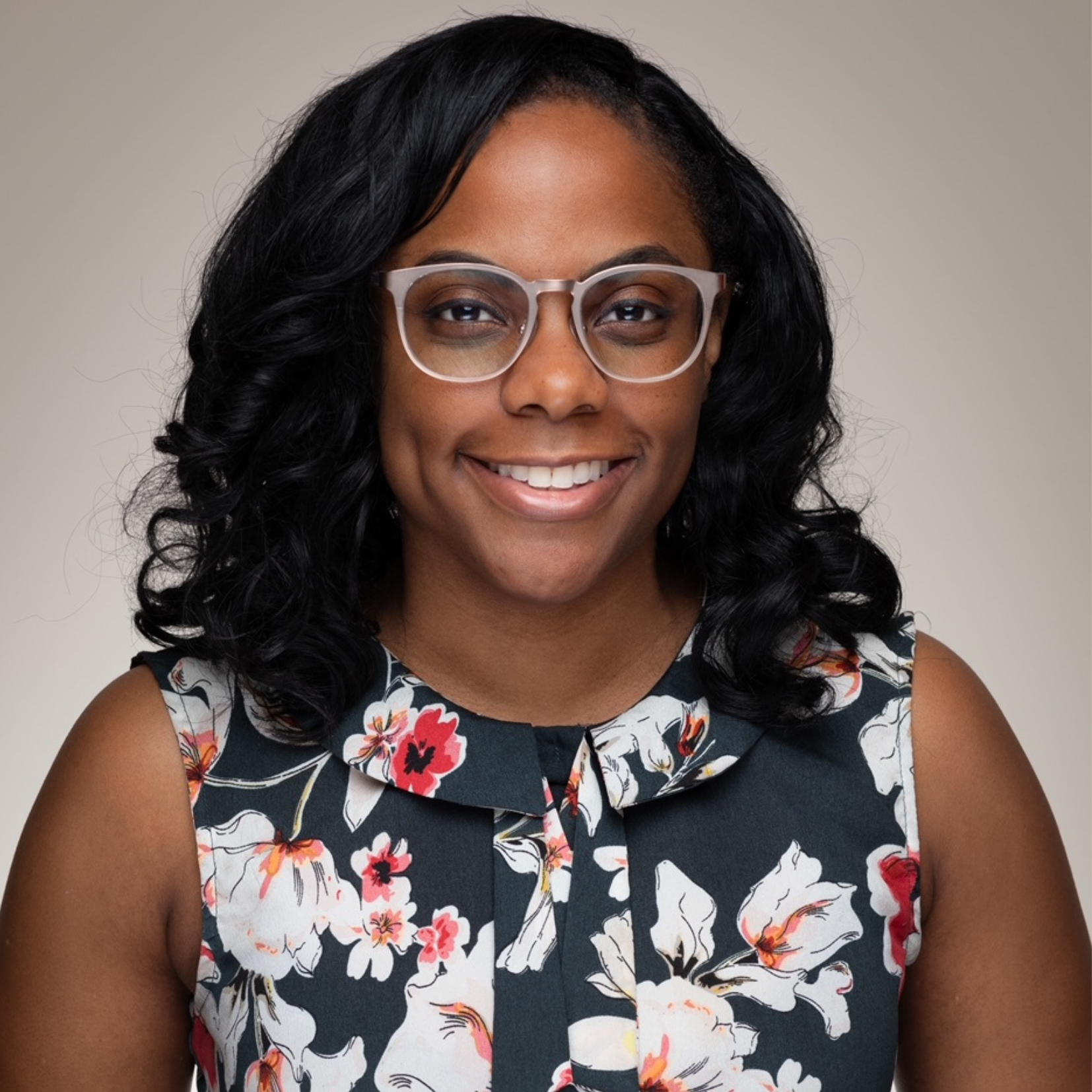 A woman with wavy black hair and clear-framed glasses smiles. She wears a sleeveless dark dress with a colorful floral pattern, standing against a neutral background.