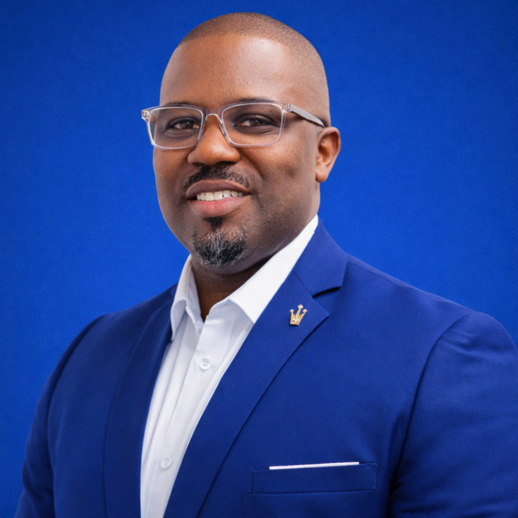 A man wearing glasses, a blue suit jacket, and a white shirt smiles in front of a solid blue background.