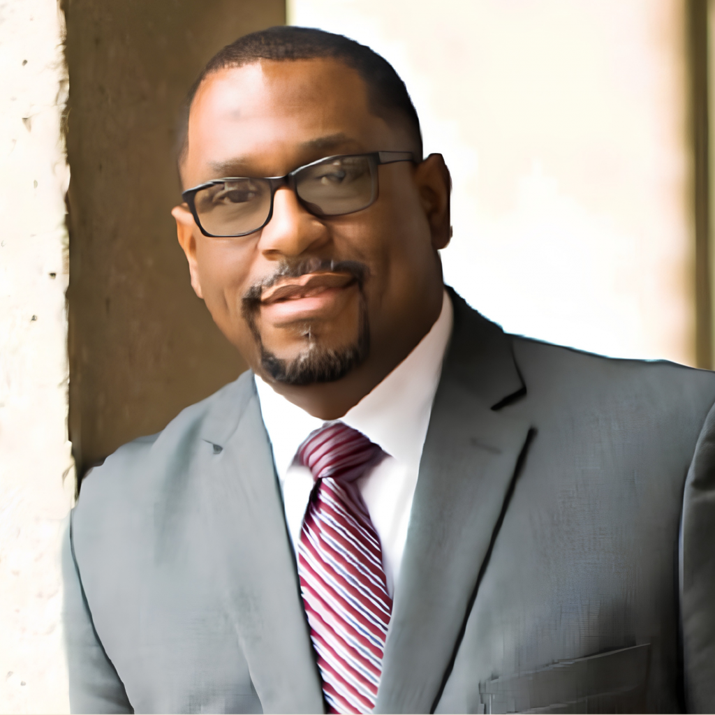 A man in a gray suit, white shirt, and red striped tie stands by a light-colored wall. He has short hair, a beard, and wears glasses, smiling slightly while looking at the camera.