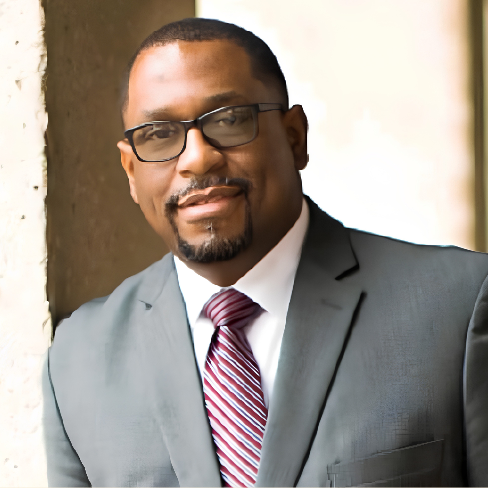 A man in a gray suit, white shirt, and red striped tie stands by a light-colored wall. He has short hair, a beard, and wears glasses, smiling slightly while looking at the camera.