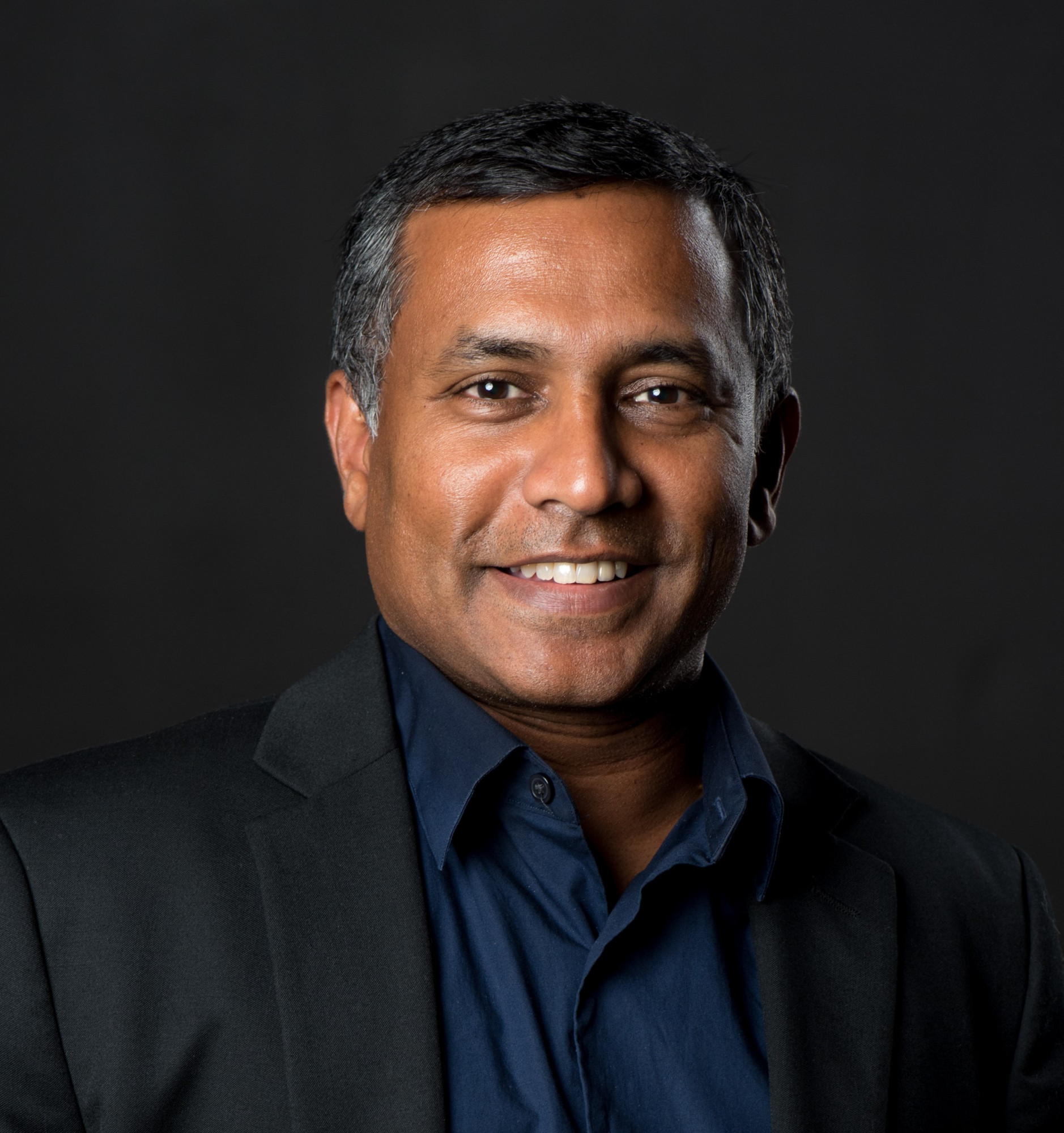 Sridhar Sunkara, a man with short dark hair, wearing a black blazer and navy shirt, smiles at the camera against a plain dark background.