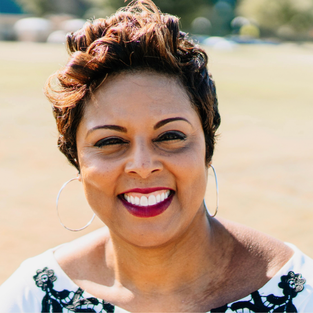 A woman with short, styled hair, large hoop earrings, and a bright smile stands outdoors in sunlight, wearing a white top with black detailing. The background is blurred greenery.