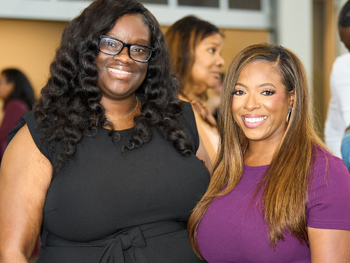 Two women smiling and standing side by side at an indoor event. Both are wearing dark dresses, and other people are blurred in the background.