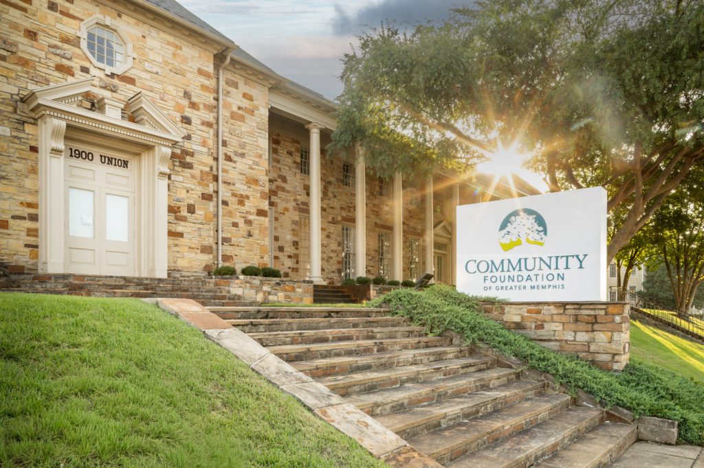 Stone building with columns and 1900 UNION above the door. Steps lead to a sign reading Community Foundation of Greater Memphis, welcoming visitors for the Grand Reopening. Sunlight filters through a tree on the right.