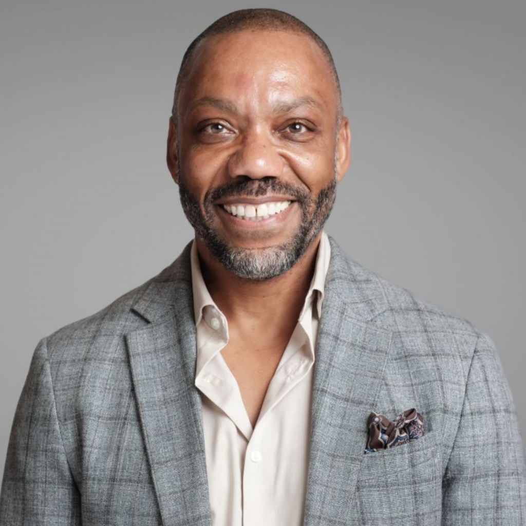 Smiling man with a short beard wearing a light gray plaid suit jacket, cream shirt, and a patterned pocket square, posing against a plain gray background.