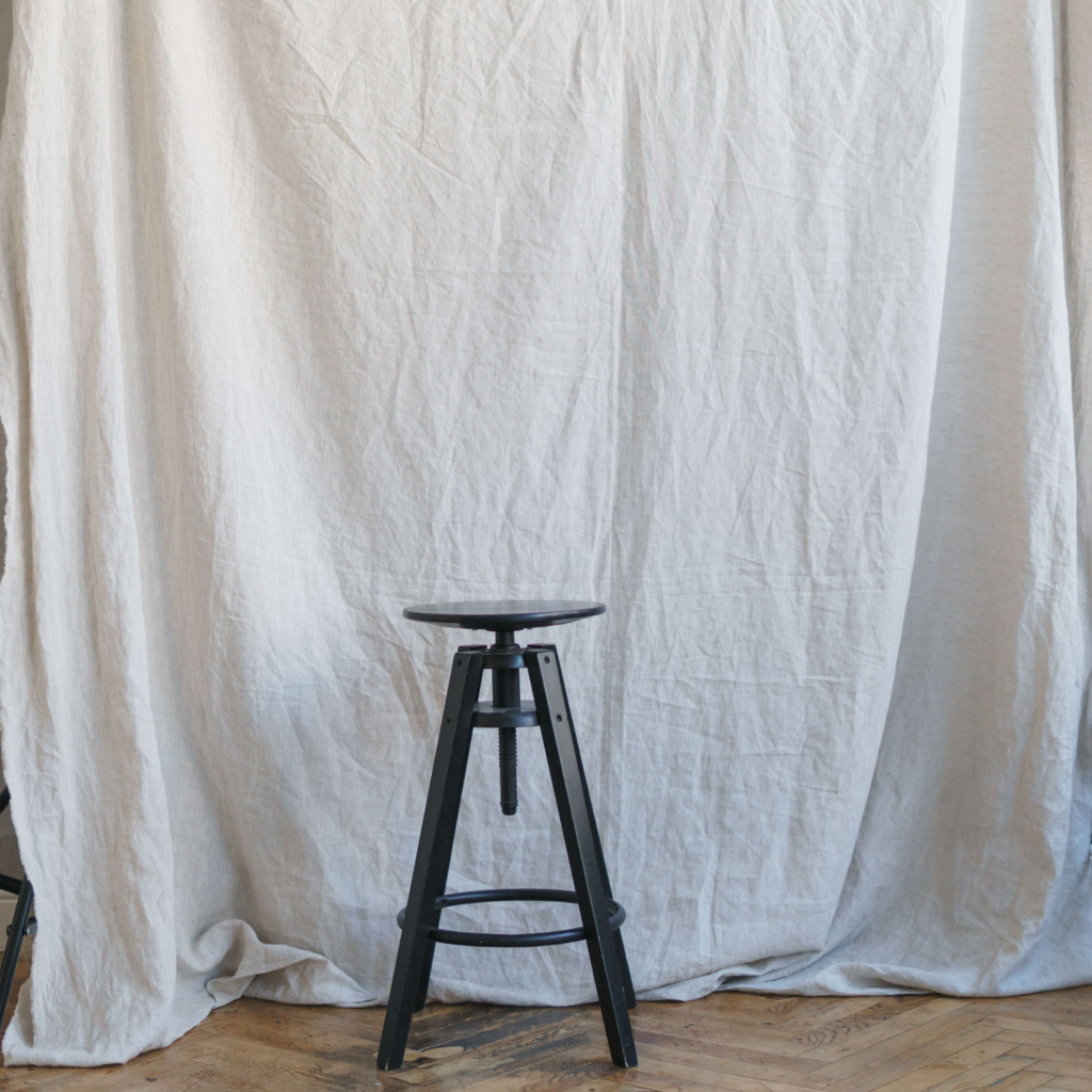 A black wooden stool stands on a wooden floor in front of a large, wrinkled white fabric backdrop, subtly informing your work with its simple elegance and versatile setting.