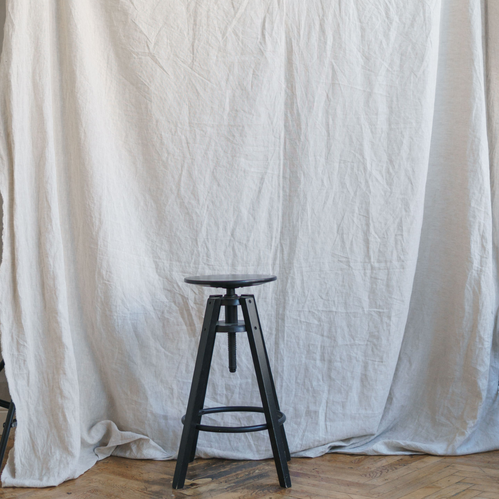 A black wooden stool stands on a wooden floor in front of a large, wrinkled white fabric backdrop, subtly informing your work with its simple elegance and versatile setting.