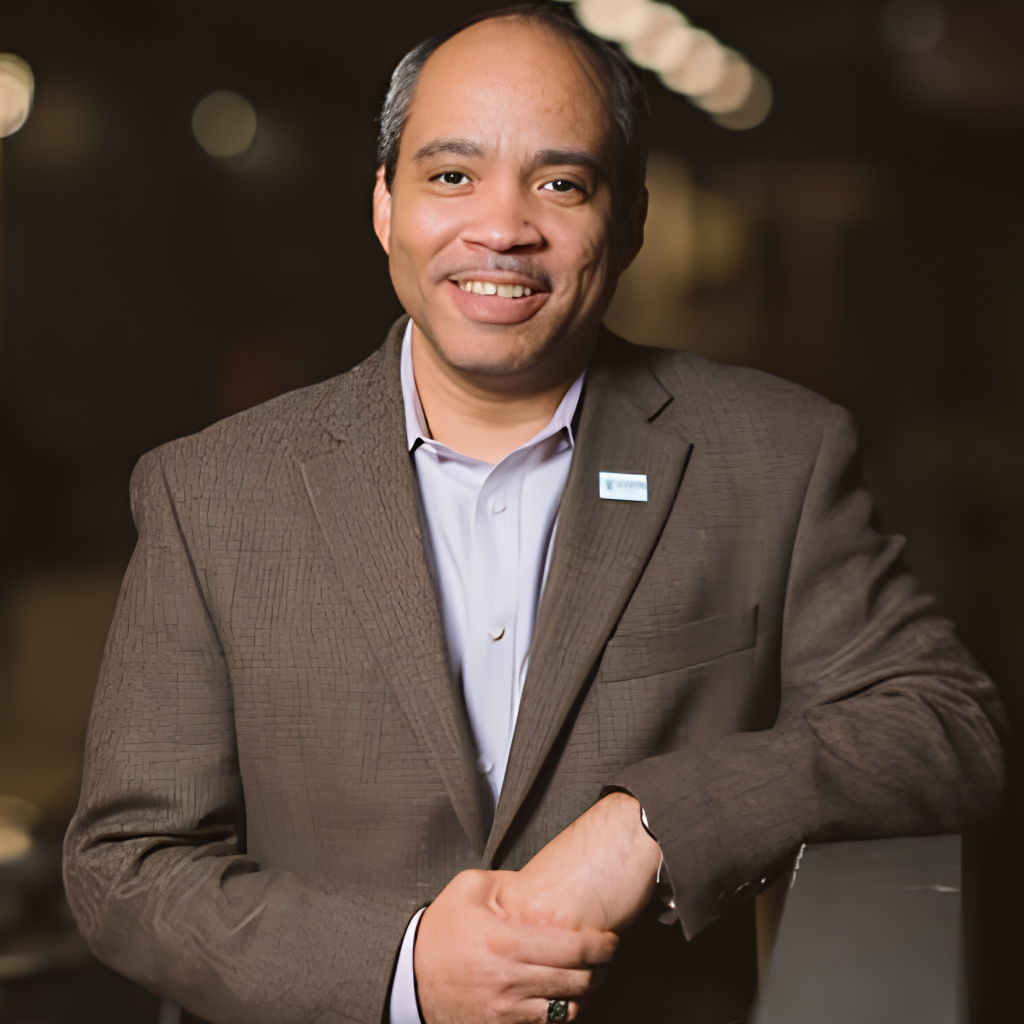 A smiling man in a brown blazer and light shirt stands casually indoors, resting one arm on a counter. The background is softly blurred, focusing attention on him.