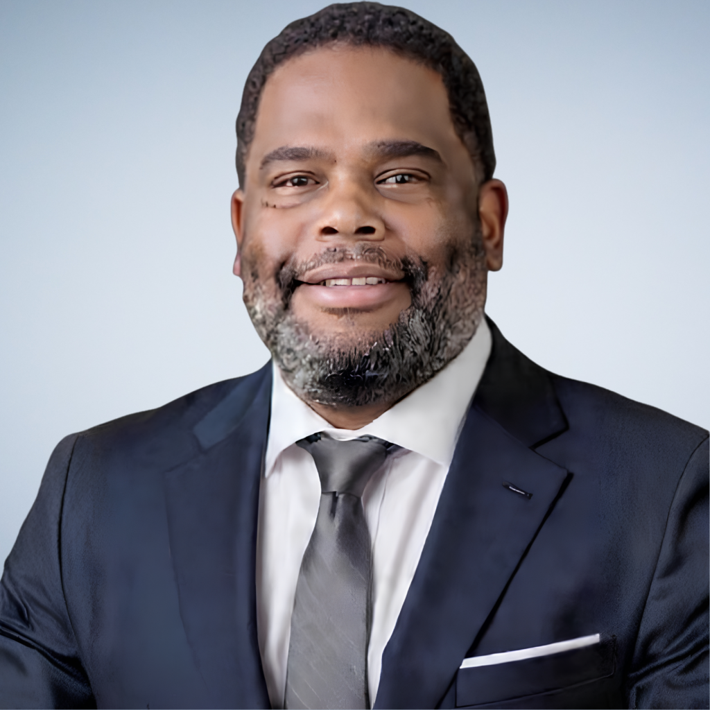 A man in a dark suit, white shirt, and gray tie smiles at the camera against a light blue background.