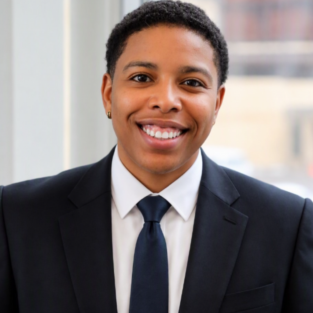 A person in a dark suit and tie smiles warmly while standing indoors near a window, with blurred buildings visible in the background.