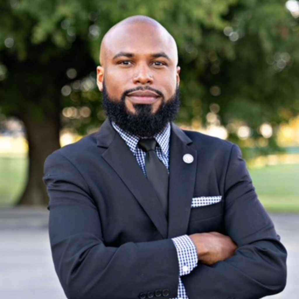 A man with a beard wearing a black suit, checked shirt, and tie stands outdoors with arms crossed, smiling confidently in front of blurred trees.
