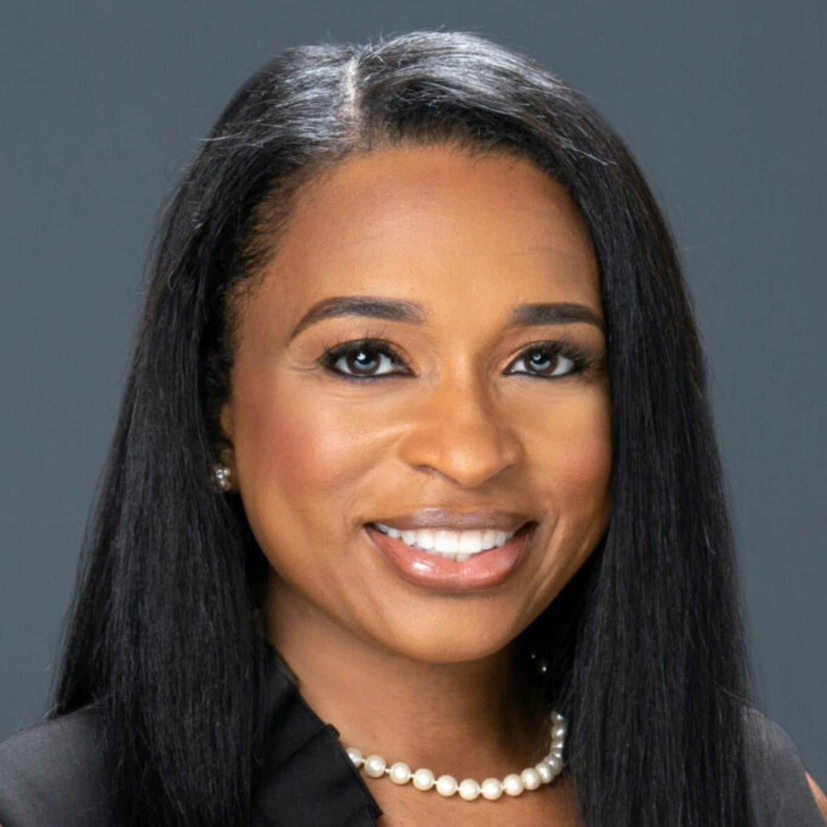 A woman with straight, dark hair, wearing a pearl necklace and a black top, smiles at the camera against a plain dark gray background.