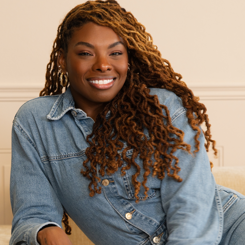 Smiling woman with long, curly, brown and blonde hair wears a denim shirt, sitting on a light couch in a softly lit room with a neutral background.