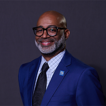 A smiling man with a bald head and gray beard, wearing glasses, a blue suit, patterned shirt, and dark tie, stands against a plain dark background. He has a blue pin on his lapel.