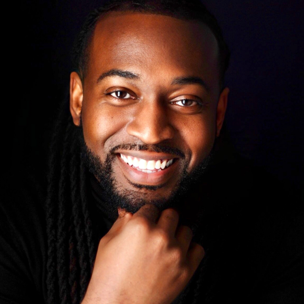 A man with long dark hair and a beard smiles warmly at the camera, resting his chin on his hand. He wears a black top and is set against a dark background.