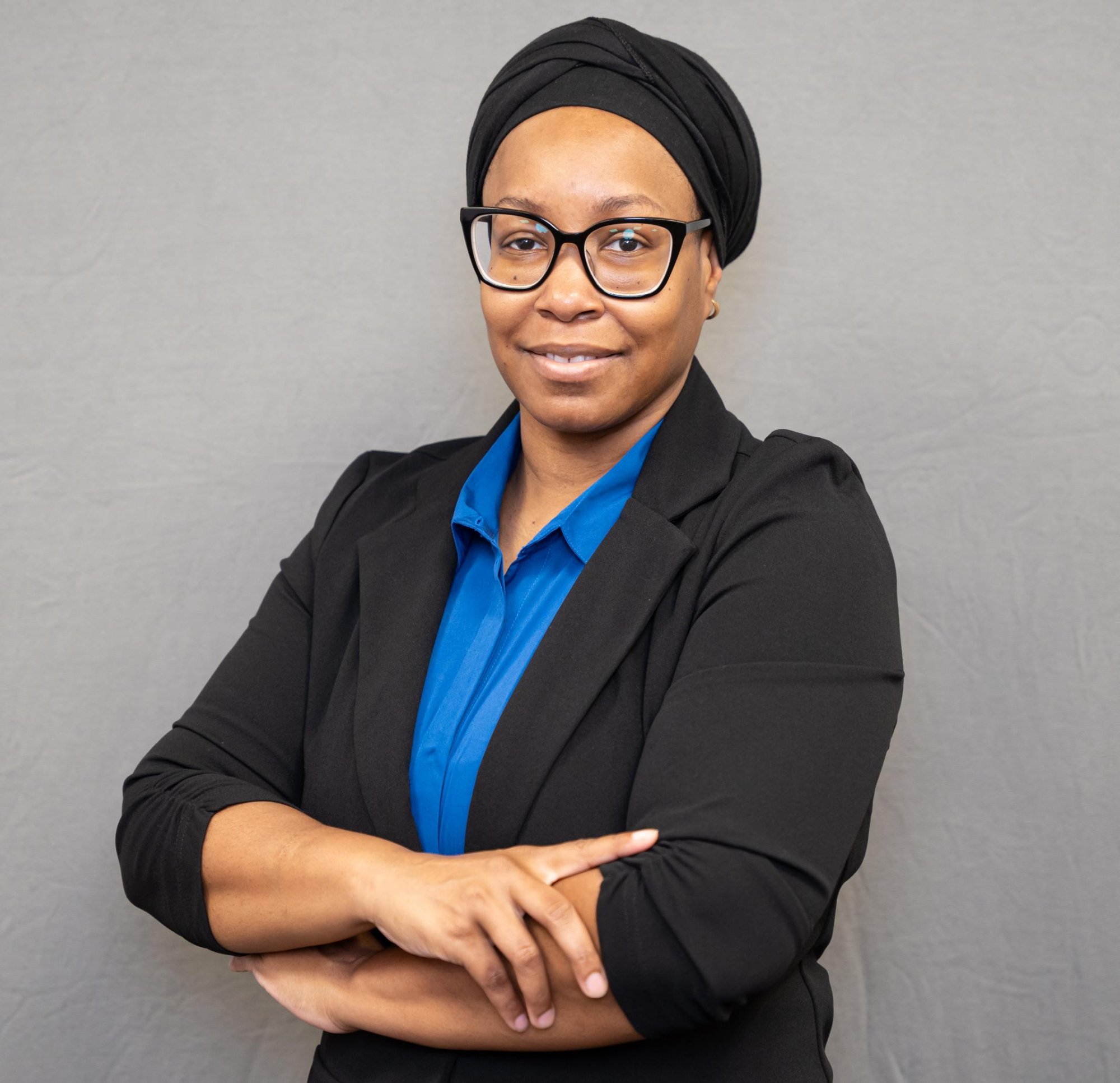 A woman wearing glasses, a black headwrap, black blazer, and blue shirt stands with arms crossed, smiling slightly in front of a plain gray background.