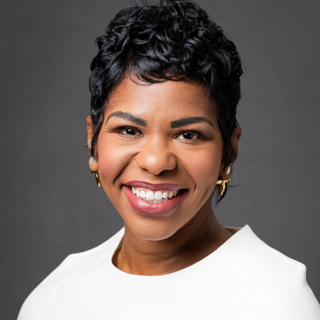 A woman with short, curly black hair smiles warmly at the camera. She is wearing gold hoop earrings and a white top, posed in front of a plain dark gray background.