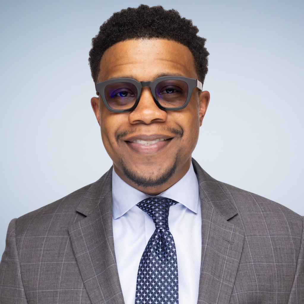 Smiling man wearing dark glasses, a gray plaid suit, a light blue shirt, and a patterned navy blue tie, standing in front of a light blue background.