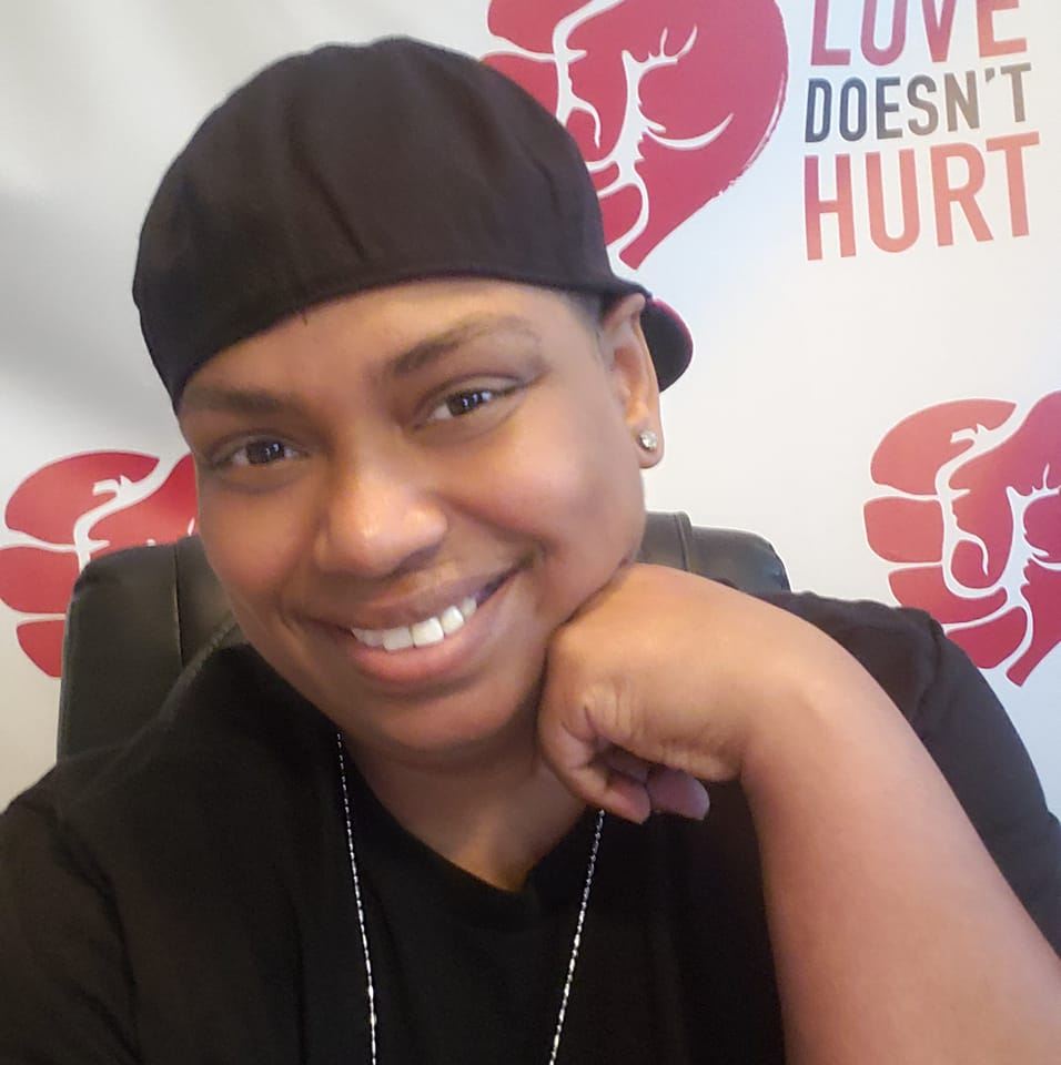Smiling person in a black shirt and cap sits with their hand on their chin, in front of a backdrop with red heart graphics and the words LOVE DOESNT HURT..