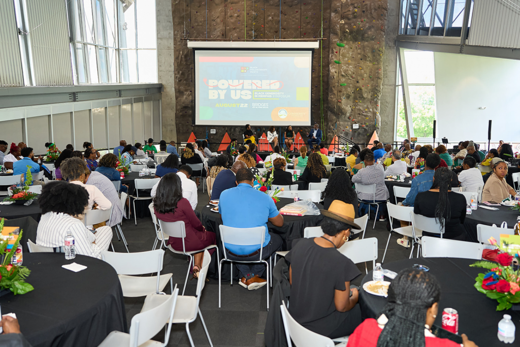 A large group of people sit at round tables during an indoor event. A panel speaks on stage near a screen displaying POWERED BY US. The room features tall windows and a climbing wall in the background.