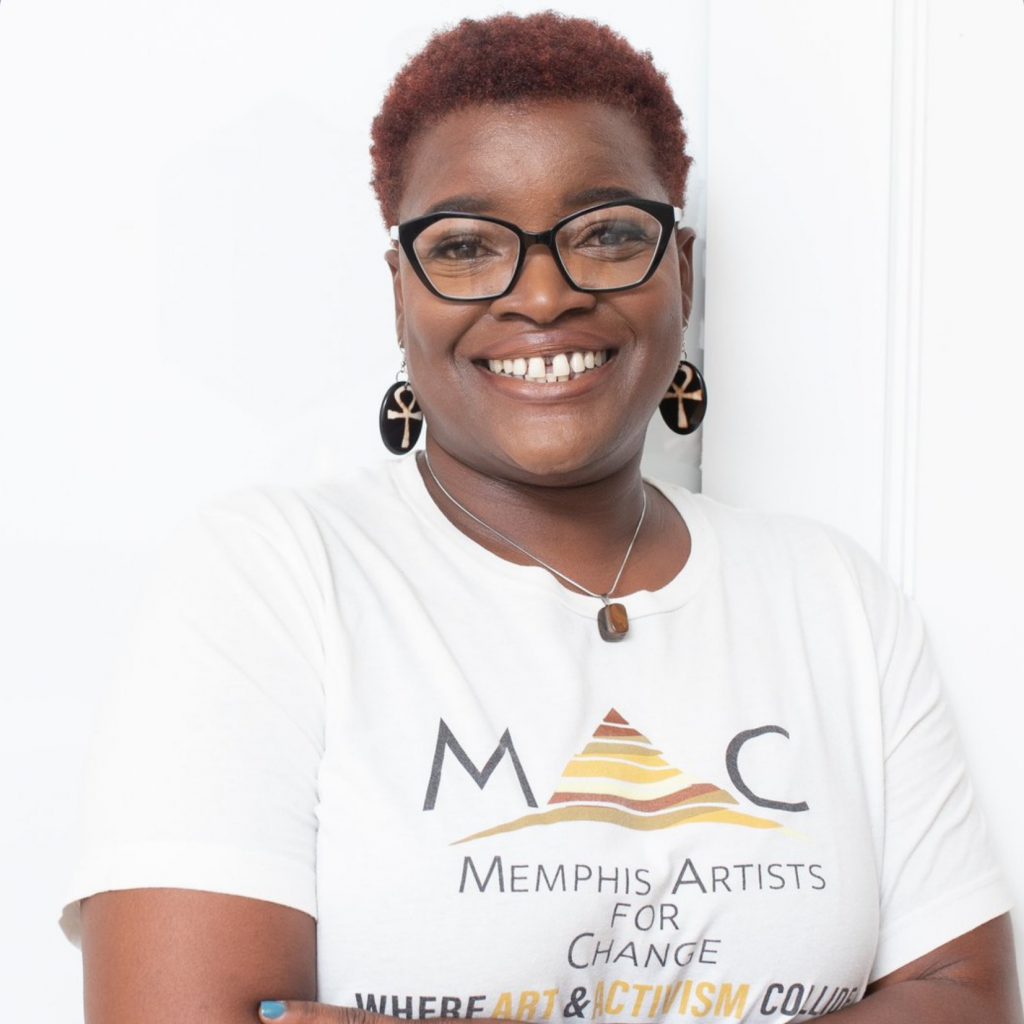 A smiling person with short hair and glasses wears earrings and a Memphis Artists for Change t-shirt, standing against a white background with arms crossed.