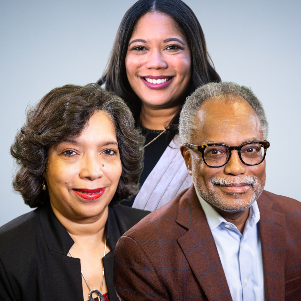 Three adults posing together in front of a light blue background; two women and one man, all smiling, wearing business attire.