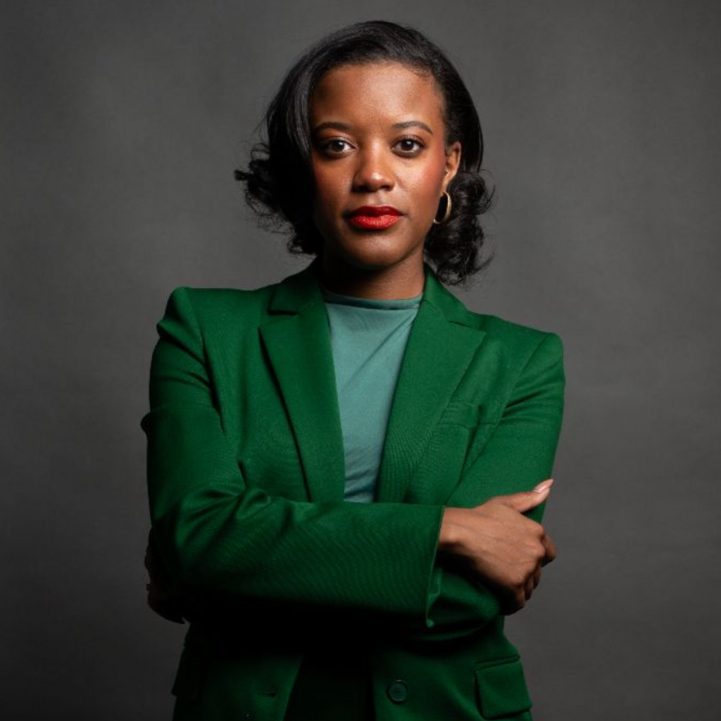 A woman in a green blazer stands with arms crossed against a dark gray background, looking confidently at the camera.