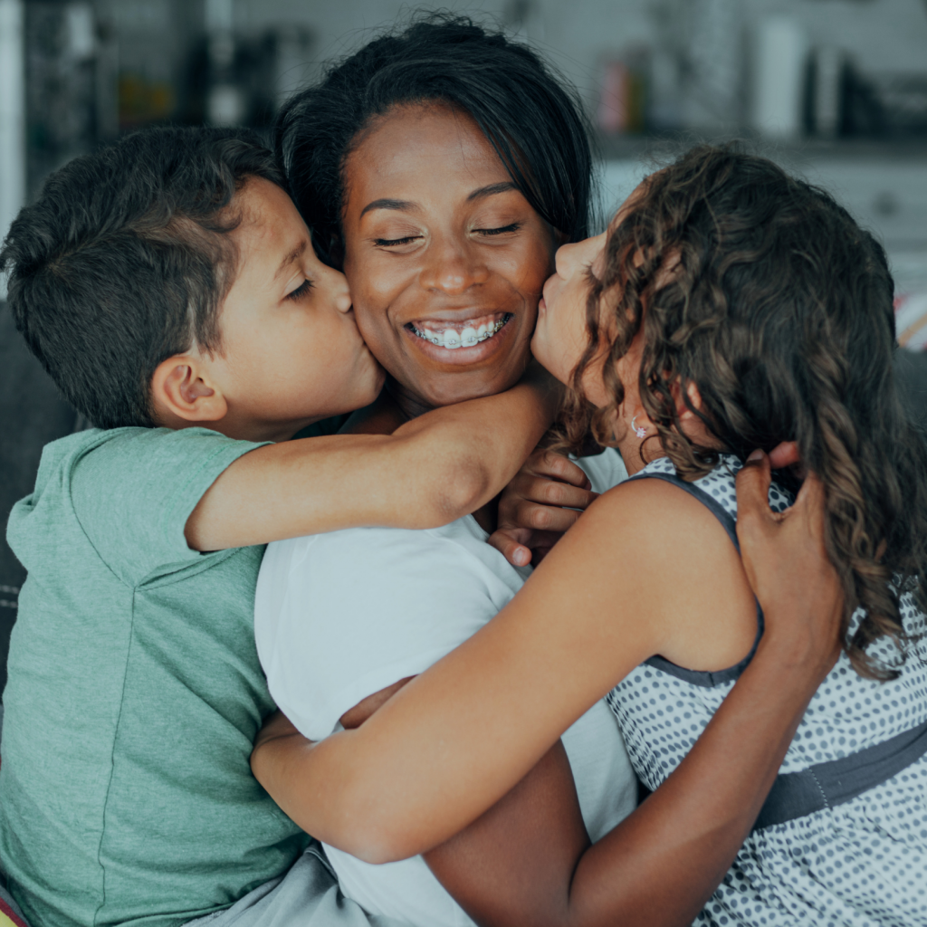 A woman smiles joyfully as two children, a boy and a girl, hug her and kiss her cheeks. They appear happy and close, sharing a loving moment together in a home setting—a beautiful reflection of building Black generational wealth through love, unity, and family support.