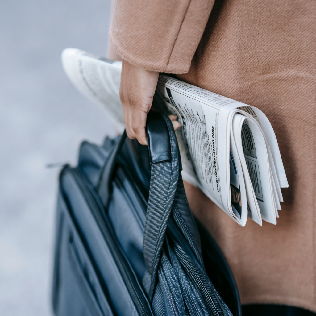 A person in a brown coat holds a black bag and a folded newspaper—certainly worth a read—under their arm, walking outside.