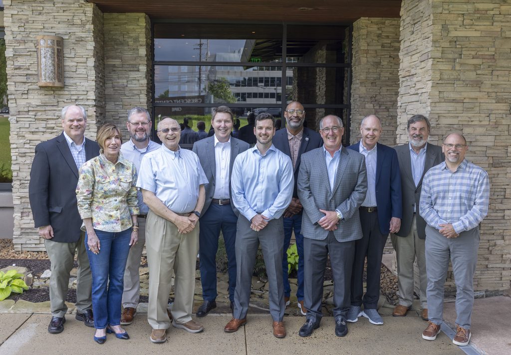 Eleven people, including two women and nine men, stand smiling in a group outside a building with stone walls and large windows. Dressed in business casual or business attire, they exude confidence and camaraderie—reflecting a team committed to investment excellence and outstanding performance.