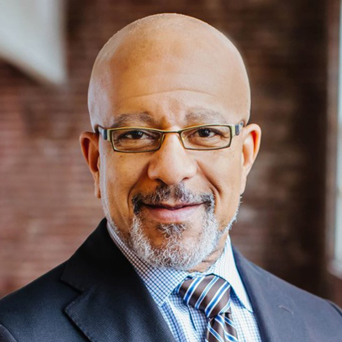 A smiling bald man with glasses, a goatee, and mustache, wearing a suit, checkered shirt, and striped tie, standing indoors with a blurred brick wall background.
