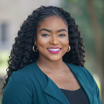 A woman with long curly hair, wearing gold hoop earrings, a teal blazer, and a necklace, smiles outdoors with a blurred natural background.