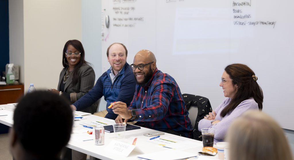Four people sit at a table in a meeting room, smiling and laughing as they discuss ideas. Notebooks, drinks, snacks, and even some notes about money are spread across the table. Notes and diagrams—including financial figures—are written on a large whiteboard behind them.