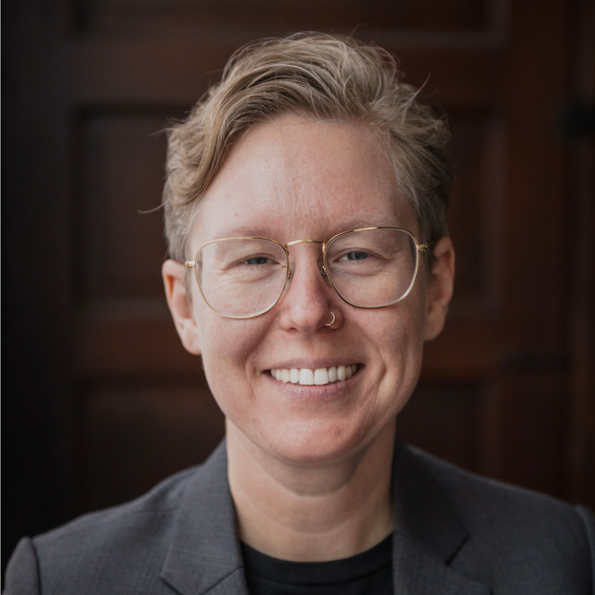Casey Bryant, a person with short, light hair, glasses, and a nose ring, smiles while wearing a dark blazer and black shirt. The background is a wooden door.