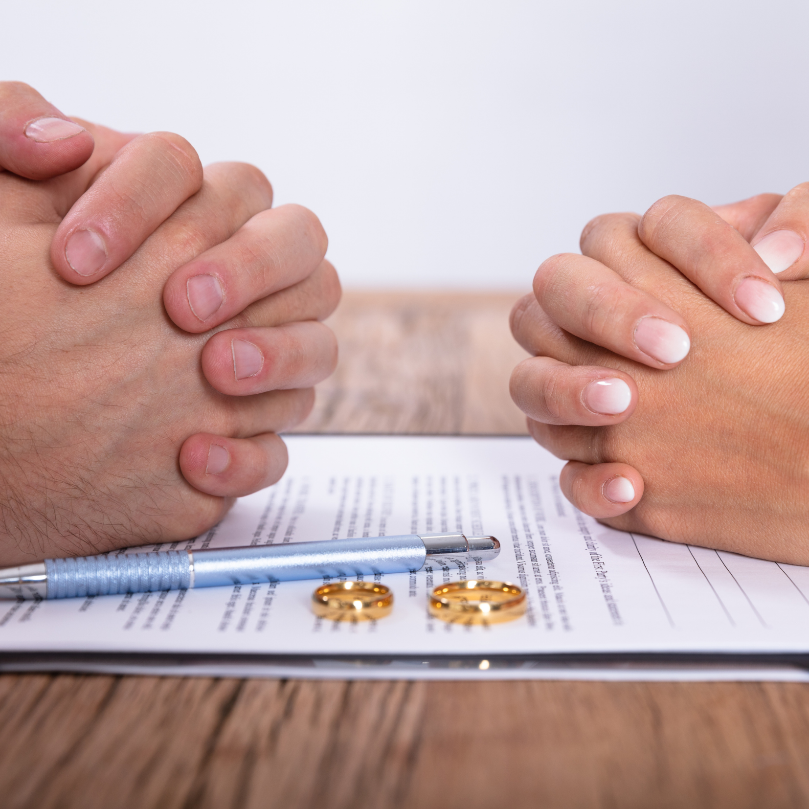 Two people sit across from each other with hands clasped over a divorce document on a table, with two gold rings and a pen placed on top.