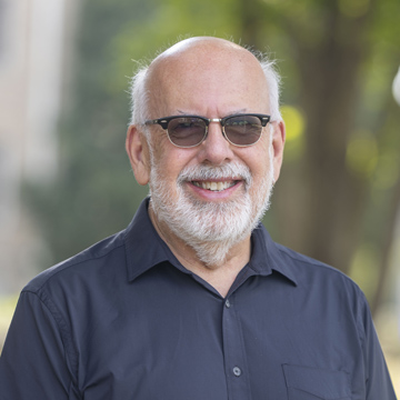 Smiling older man with a white beard and glasses wearing a dark shirt, standing outdoors with trees blurred in the background.