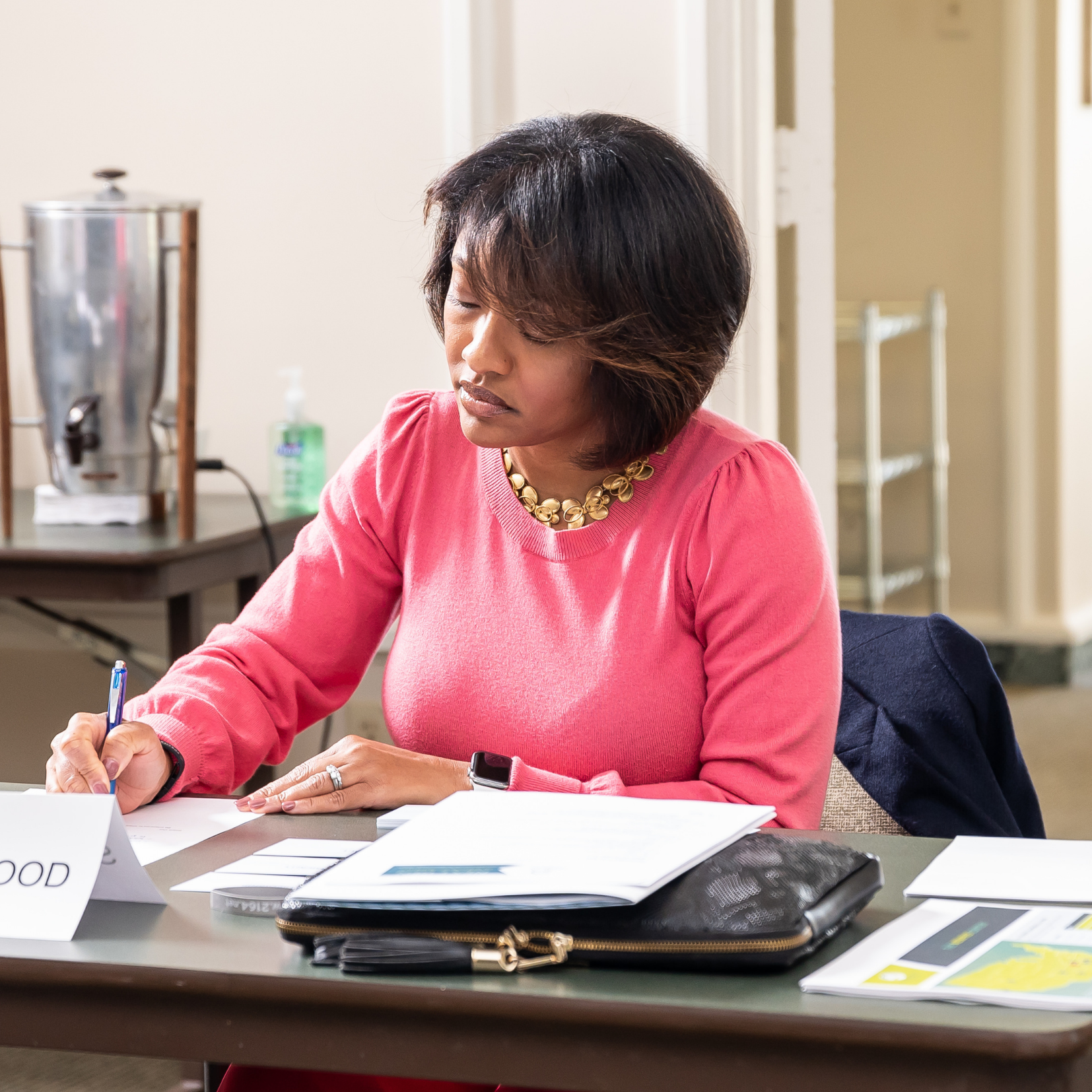 A woman in a pink top writes at a desk with papers, a folder, and a nameplate. A coffee dispenser and hand sanitizer are in the background.