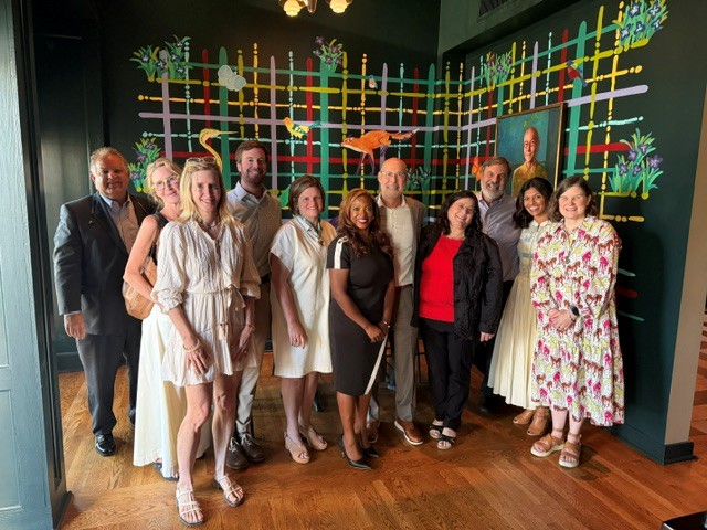A group of twelve adults, including a philanthropic advisor, stands together indoors on a wood floor, smiling for a group photo in front of a colorful grid-patterned wall mural featuring animals and plants.