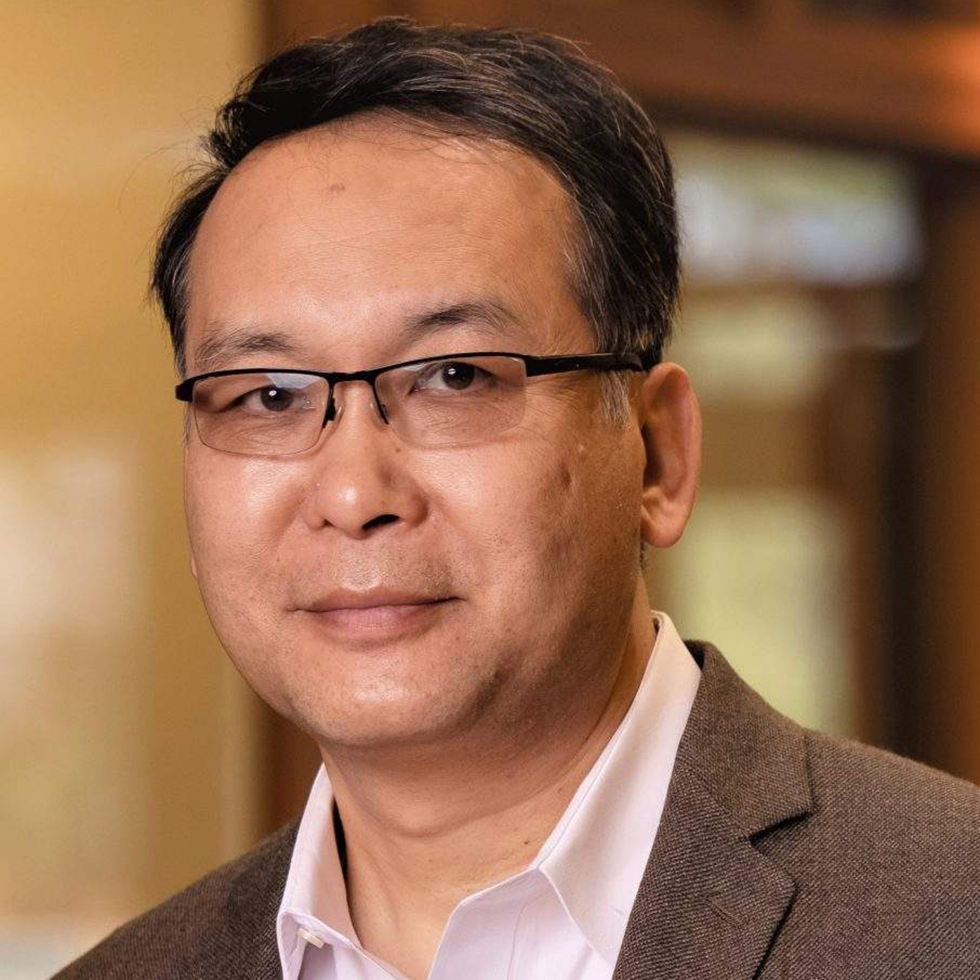 A man wearing glasses and a brown blazer over a white shirt, standing indoors with a soft, blurred background.