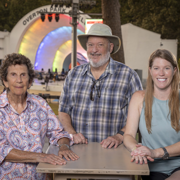 Three people stand smiling at a table outdoors, with a large band shell and colorful lights in the background. Rooted in the vibrant spirit of Memphis, trees and people can be seen behind them at what appears to be a lively park event.