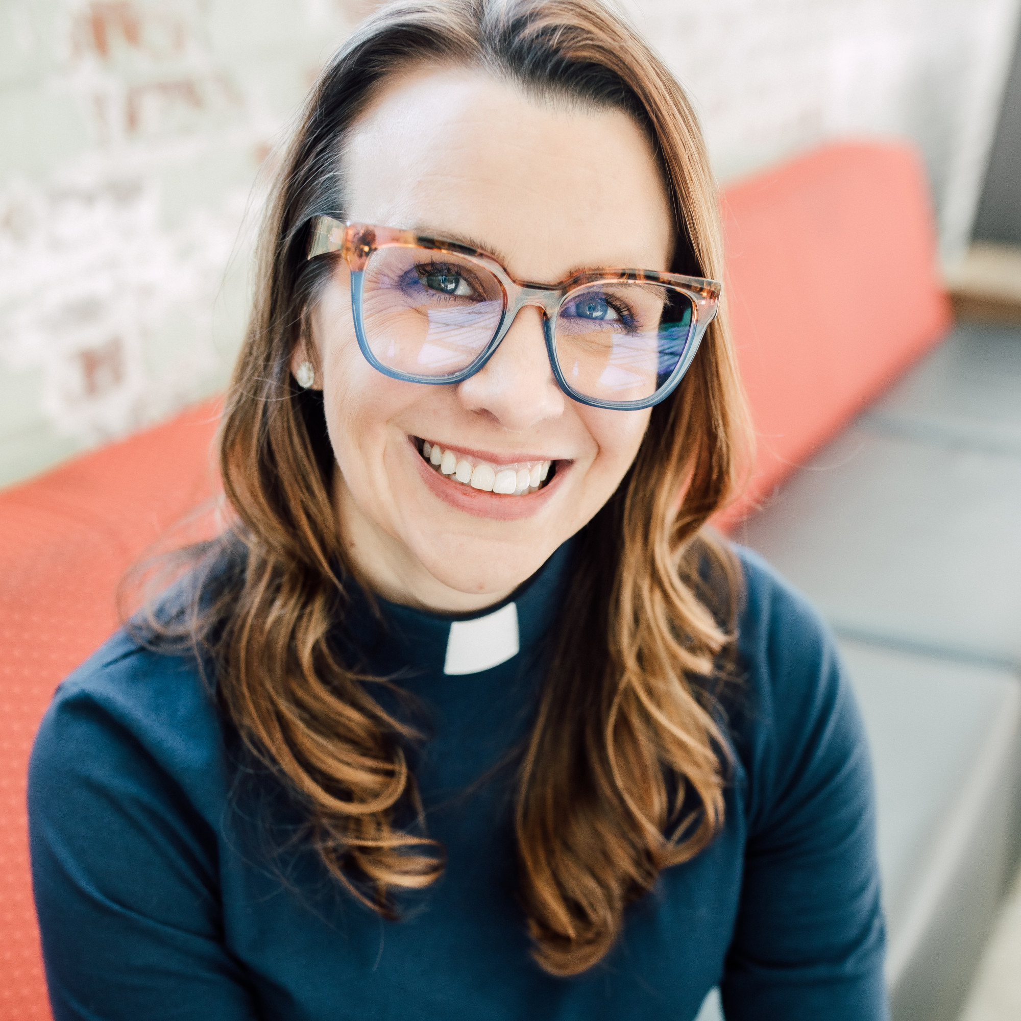A woman with long brown hair, wearing large glasses and a clerical collar, smiles while sitting on a red-cushioned bench against a light-colored, exposed brick wall; she is known as Casey Bryant.
