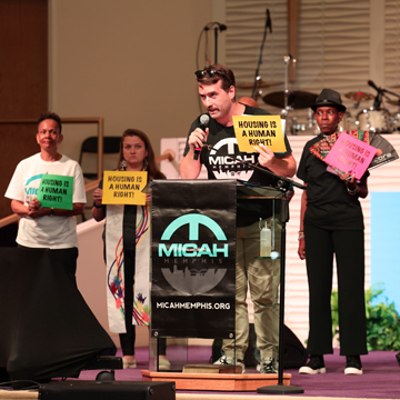 A man speaks at a podium labeled MICAH Memphis while three people behind him hold signs saying Housing is a human right and Housing is a vital human right, emphasizing the importance of playing the long game to secure lasting change for their community.