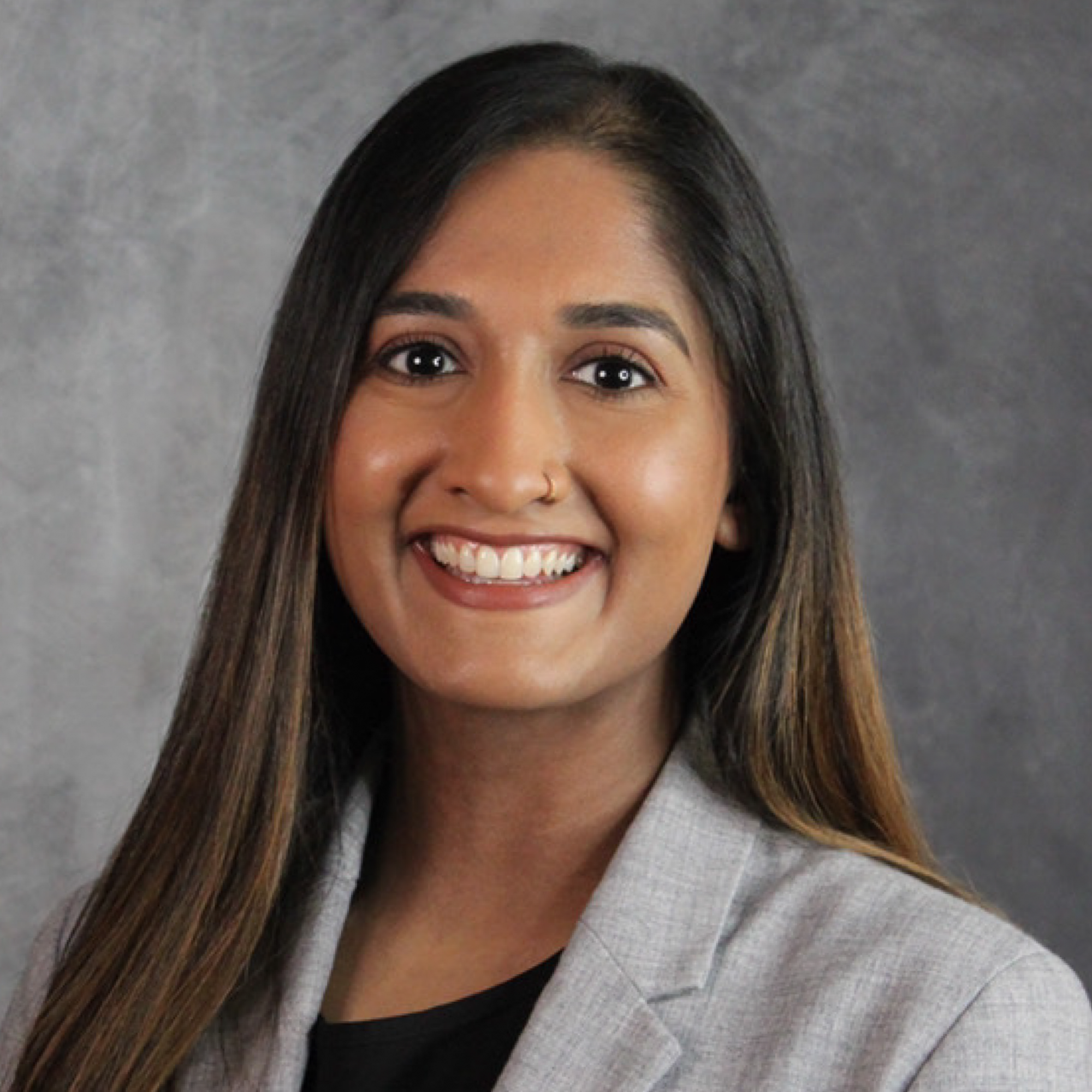 Casey Bryant, a woman with long, straight brown hair, smiles at the camera. She is wearing a light gray blazer over a black top, with a gray mottled background behind her.