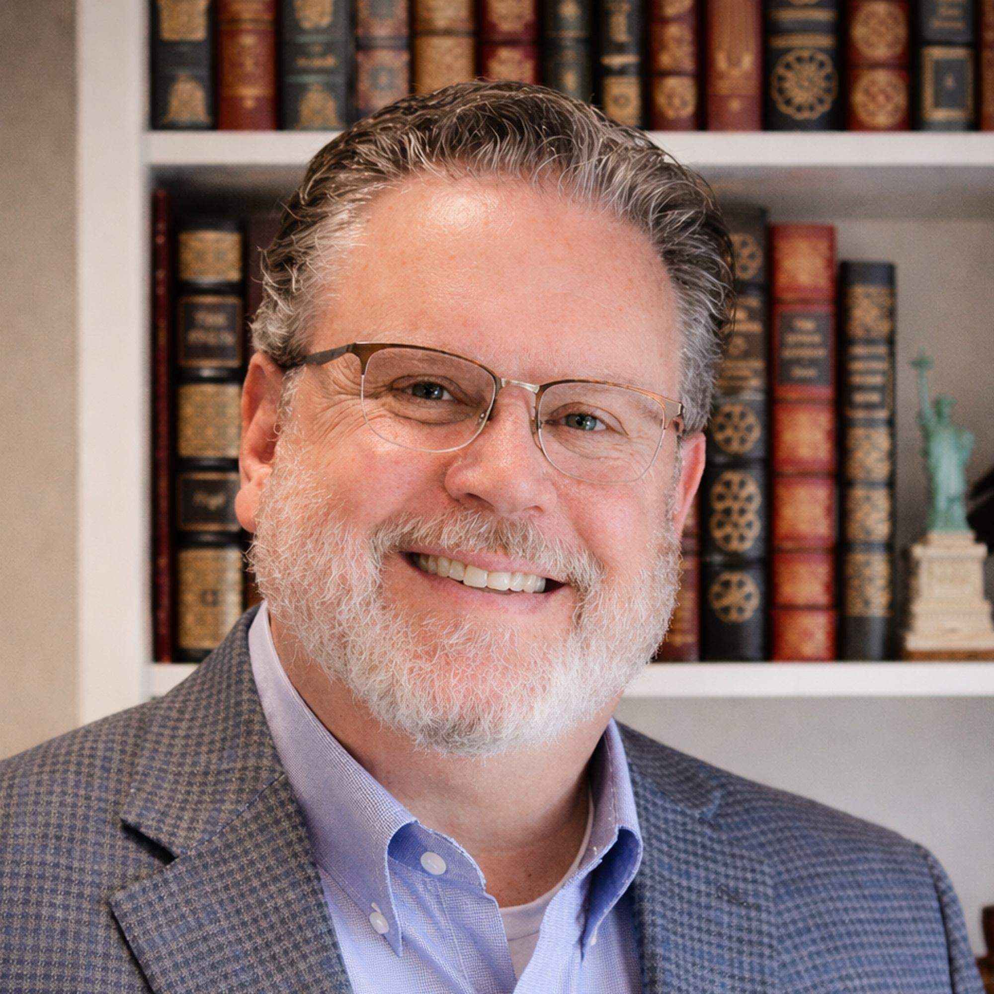 Smiling man with glasses and a gray beard, Casey Bryant is wearing a blue checkered blazer and collared shirt, standing in front of a bookshelf with colorful books and a small Statue of Liberty figurine.