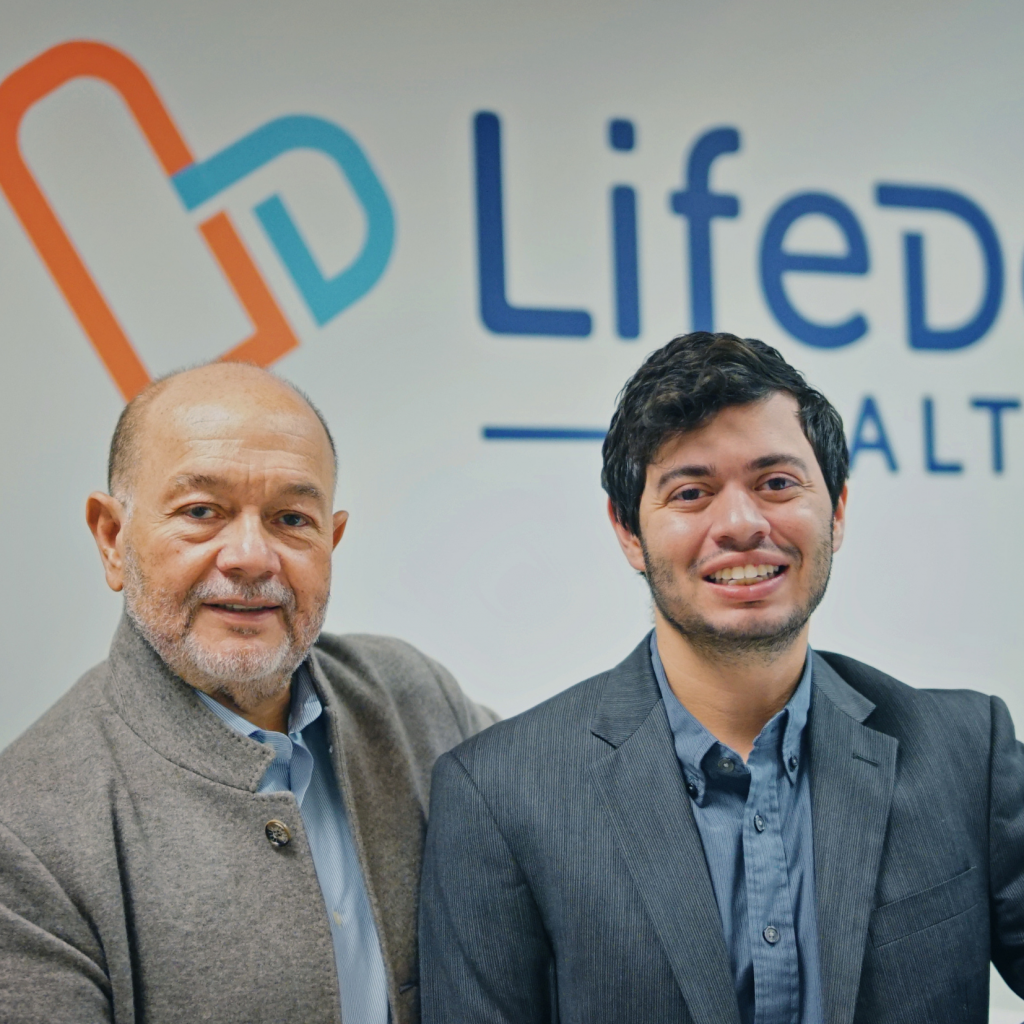 Two men in business attire stand smiling in front of a wall with the LifeDe logo and partial word HEALTH visible, celebrating Latinx Leadership during National Hispanic Heritage Month.