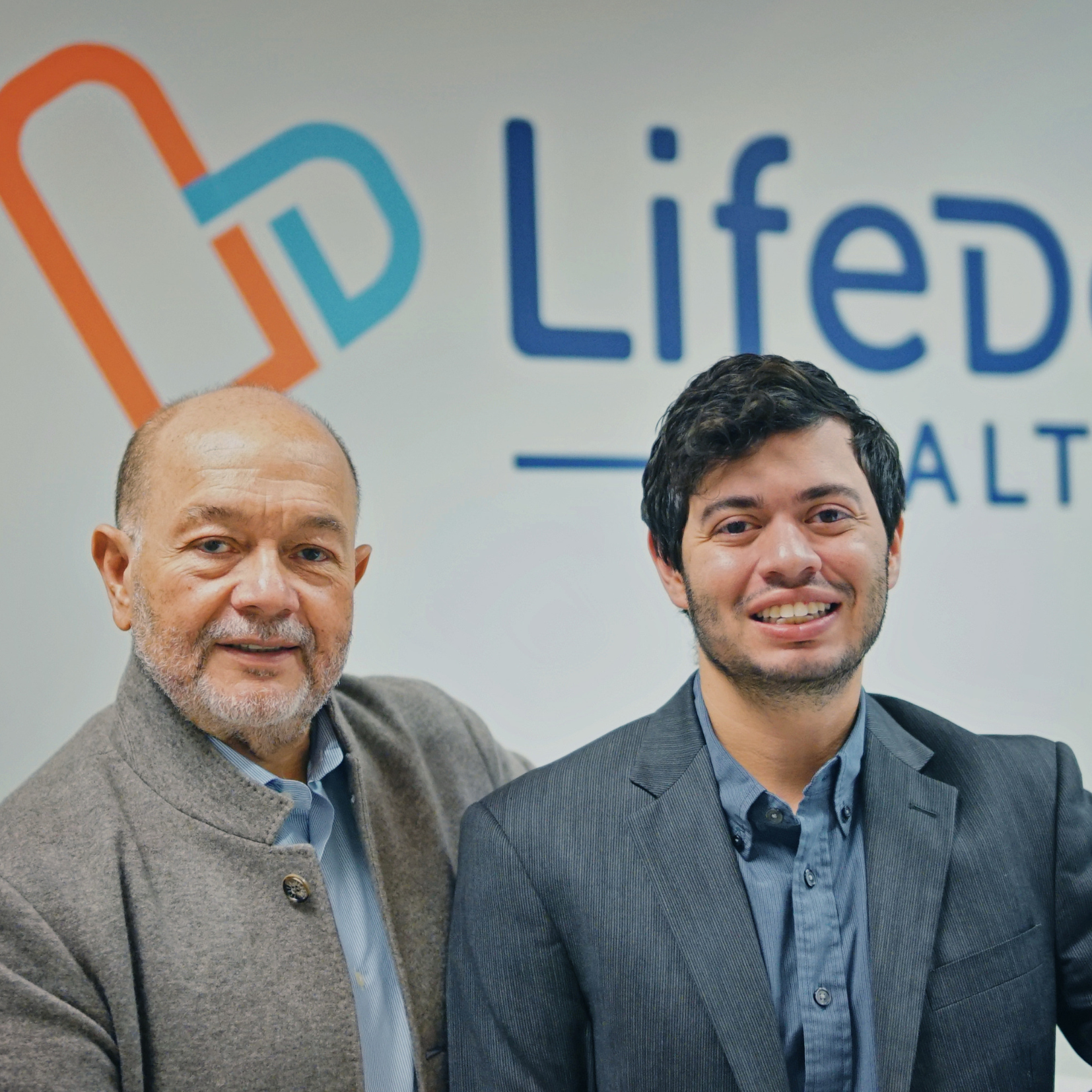 Two men in business attire stand smiling in front of a wall with the LifeDe logo and partial word HEALTH visible, celebrating Latinx Leadership during National Hispanic Heritage Month.