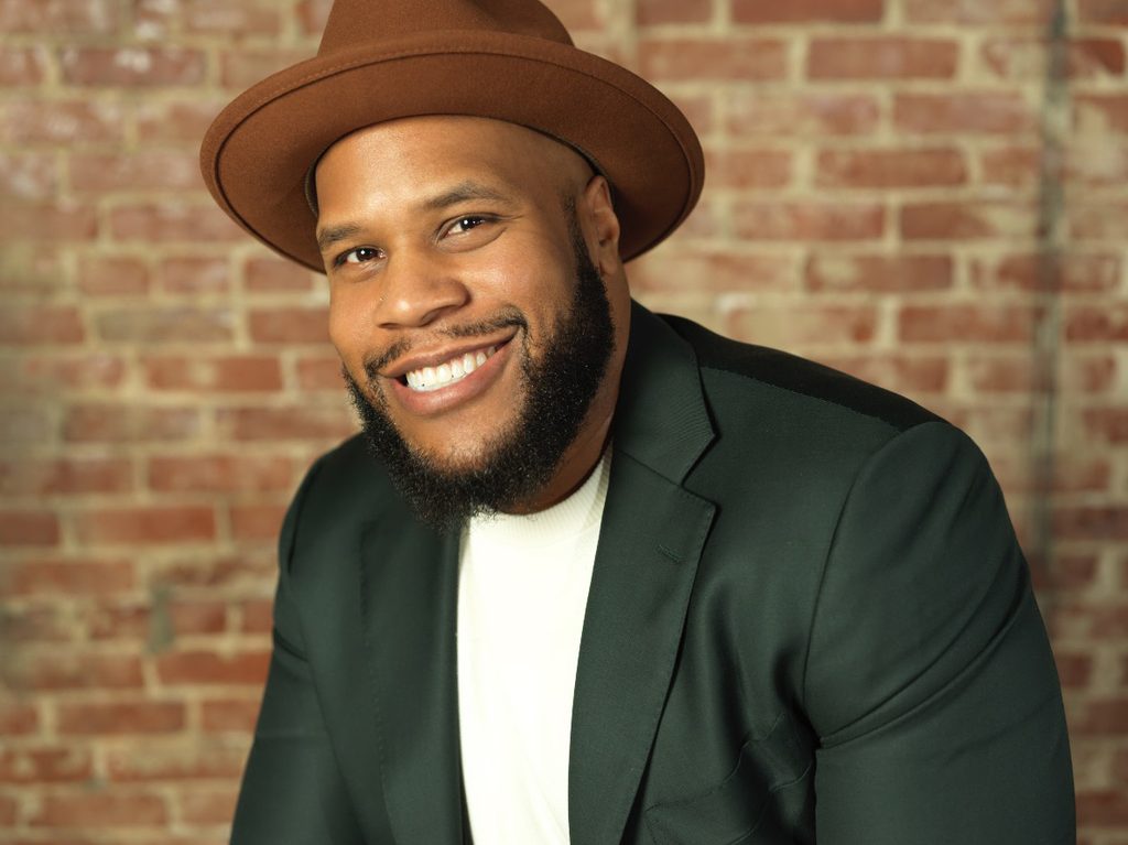 A man wearing a brown hat, dark green blazer, and white shirt smiles in front of a brick wall background, exuding the confidence of someone who knows how to handle money.